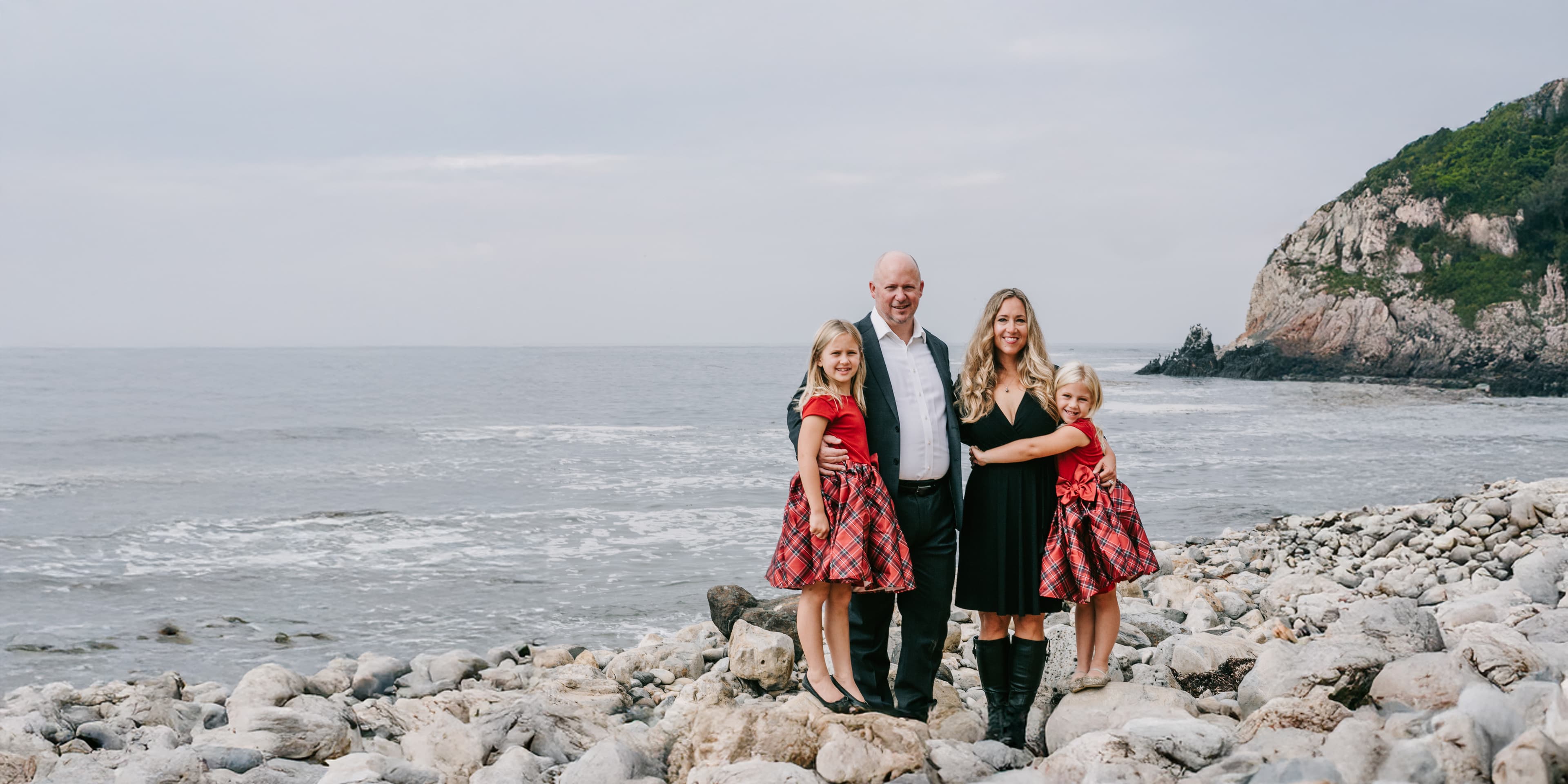 A family of 4 poses for pictures on a rocky shore, with ocean waves and a mossy cliff behind them.