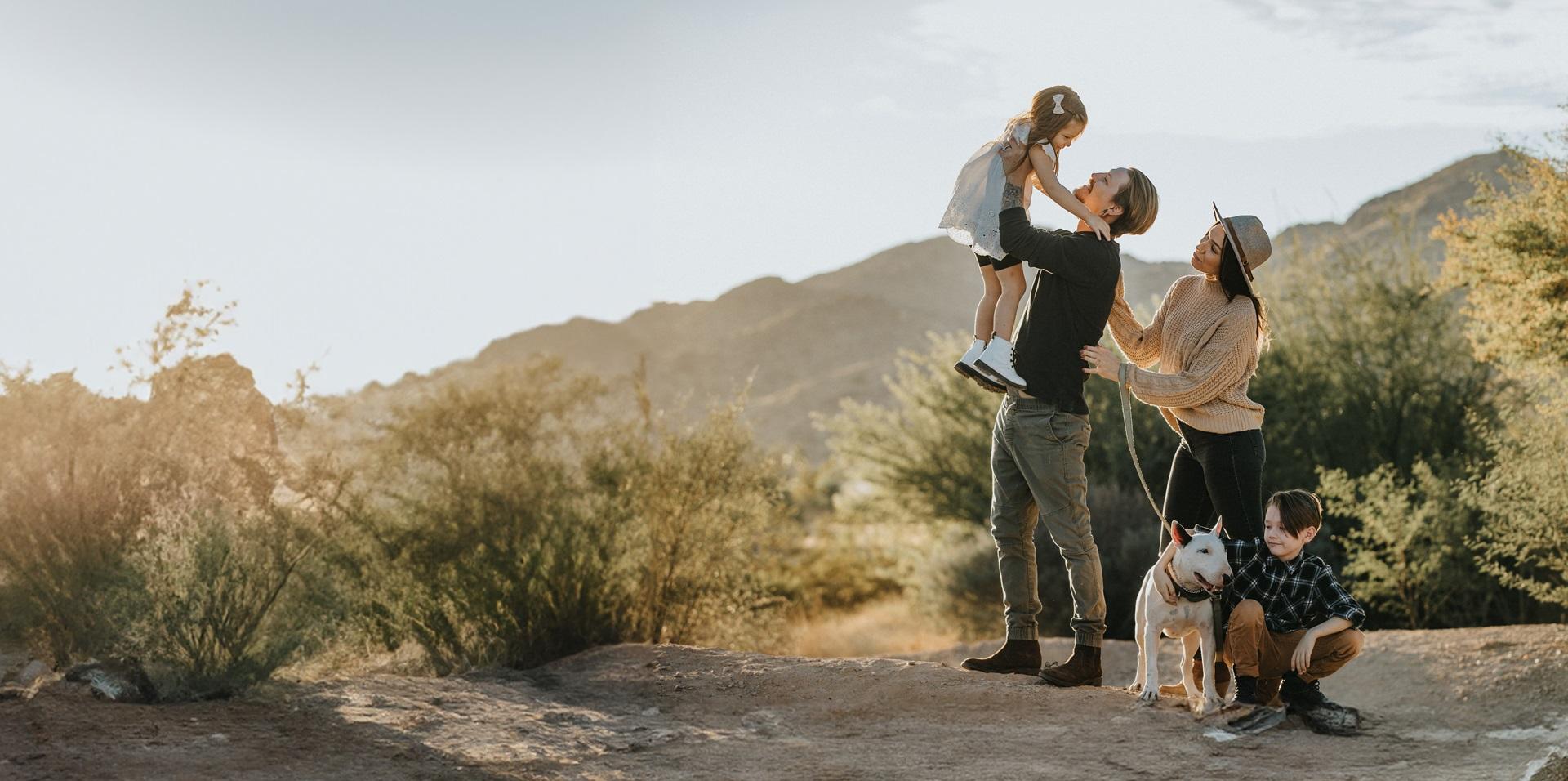 A family stands in the desert with their dog, surrounded by sandy dunes and a clear blue sky