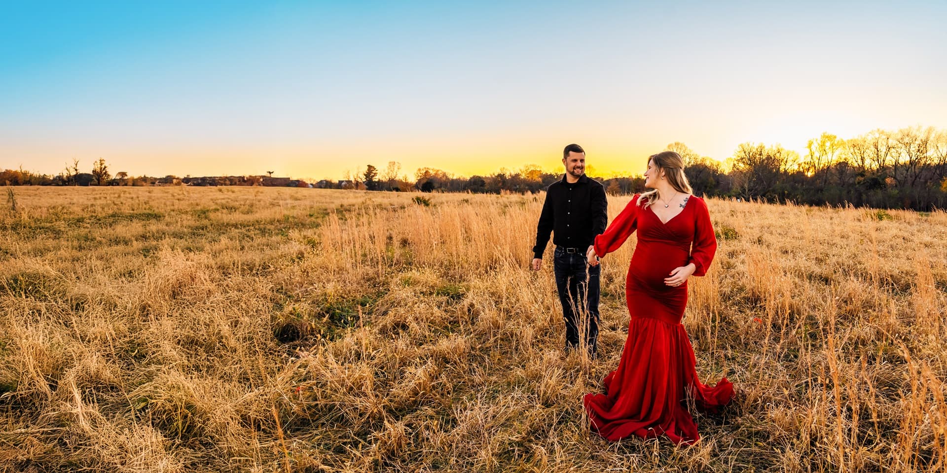 A couple walks hand in hand in a lush field smiling at each other