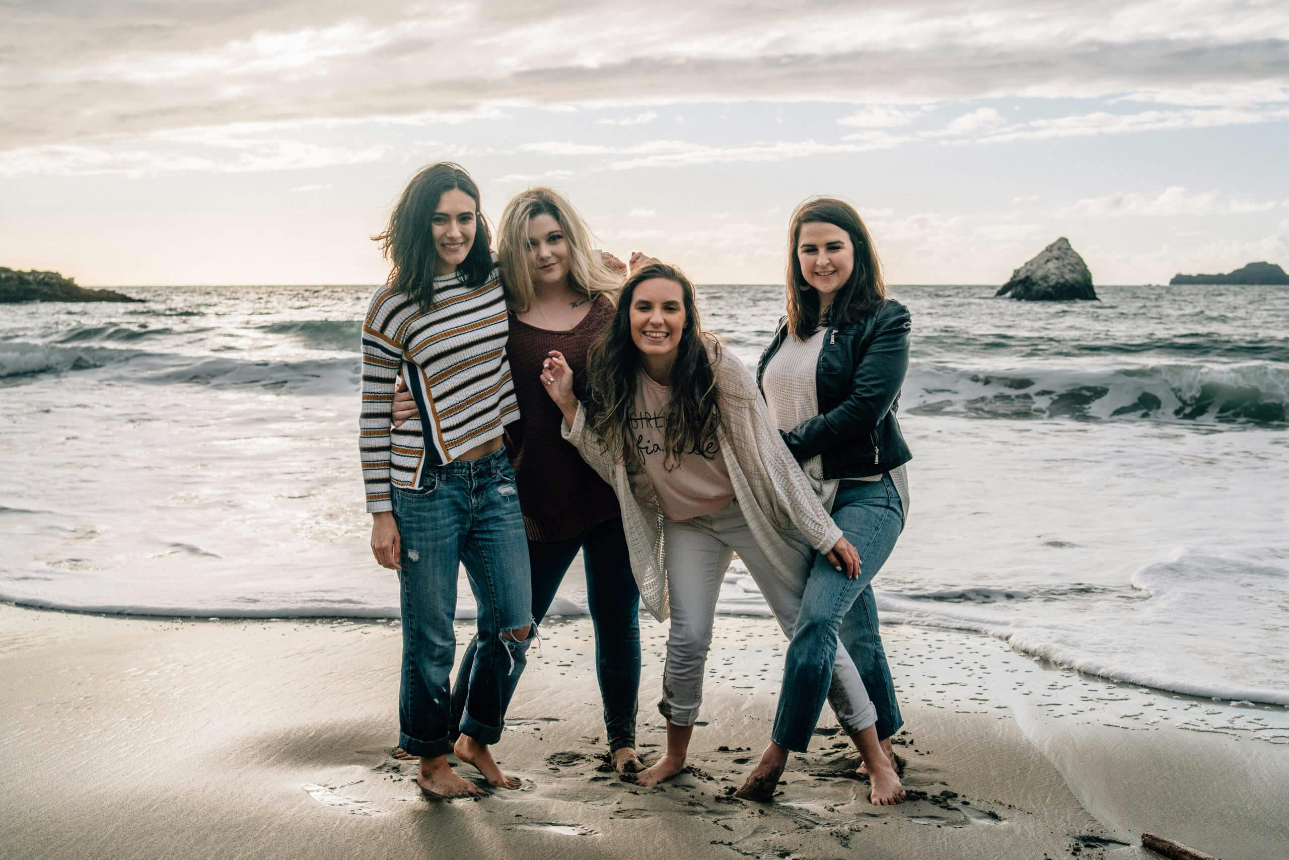 Four women standing on a beach with waves and rocks in the background.