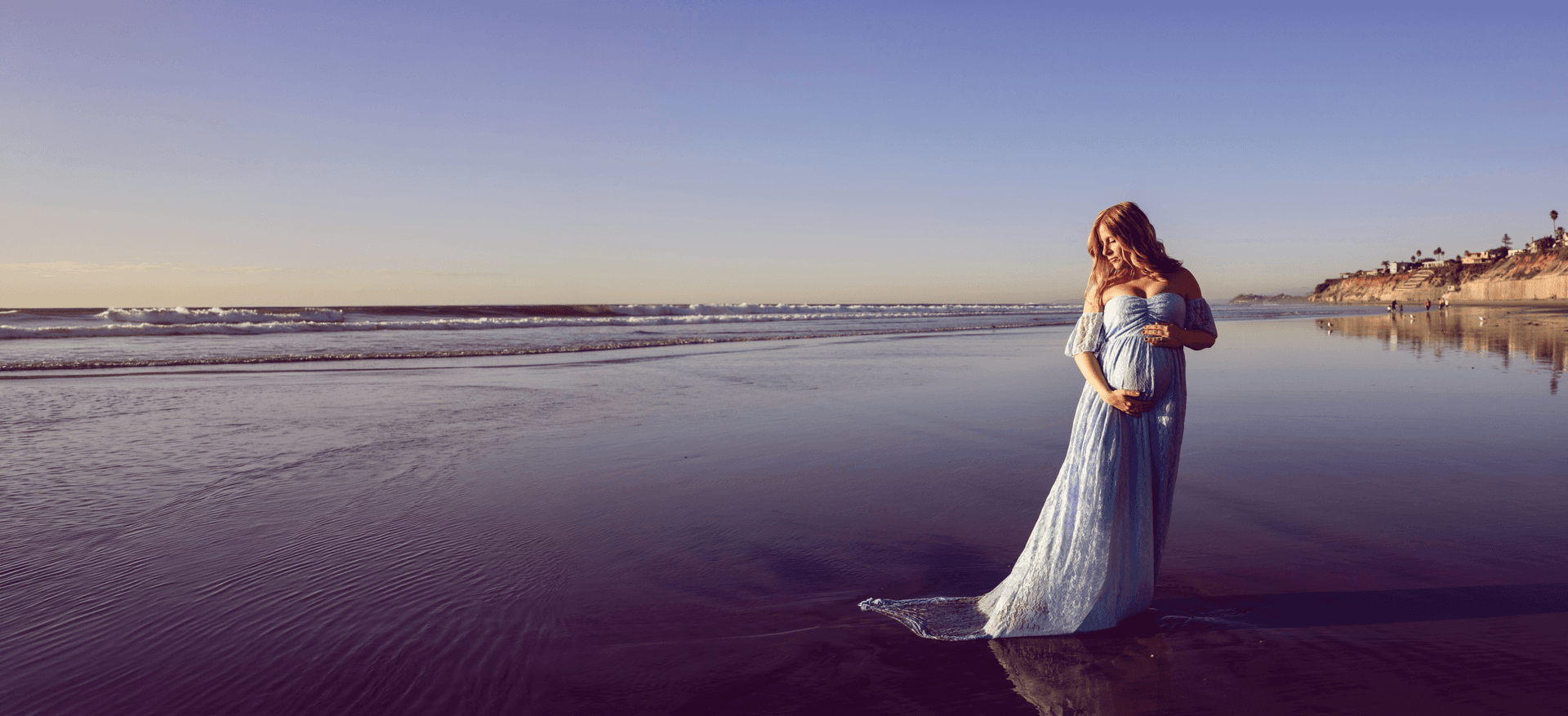 Expecting mother in a flowing blue gown standing on a beach at sunset