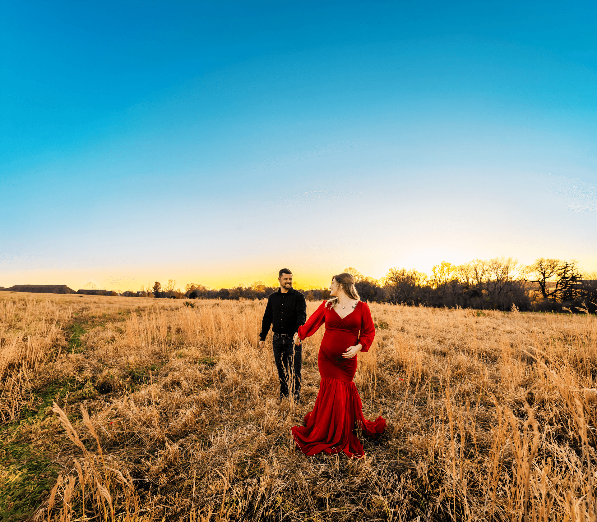 Expecting couple walking hand in hand through a golden field at sunset, woman in a flowing red dress