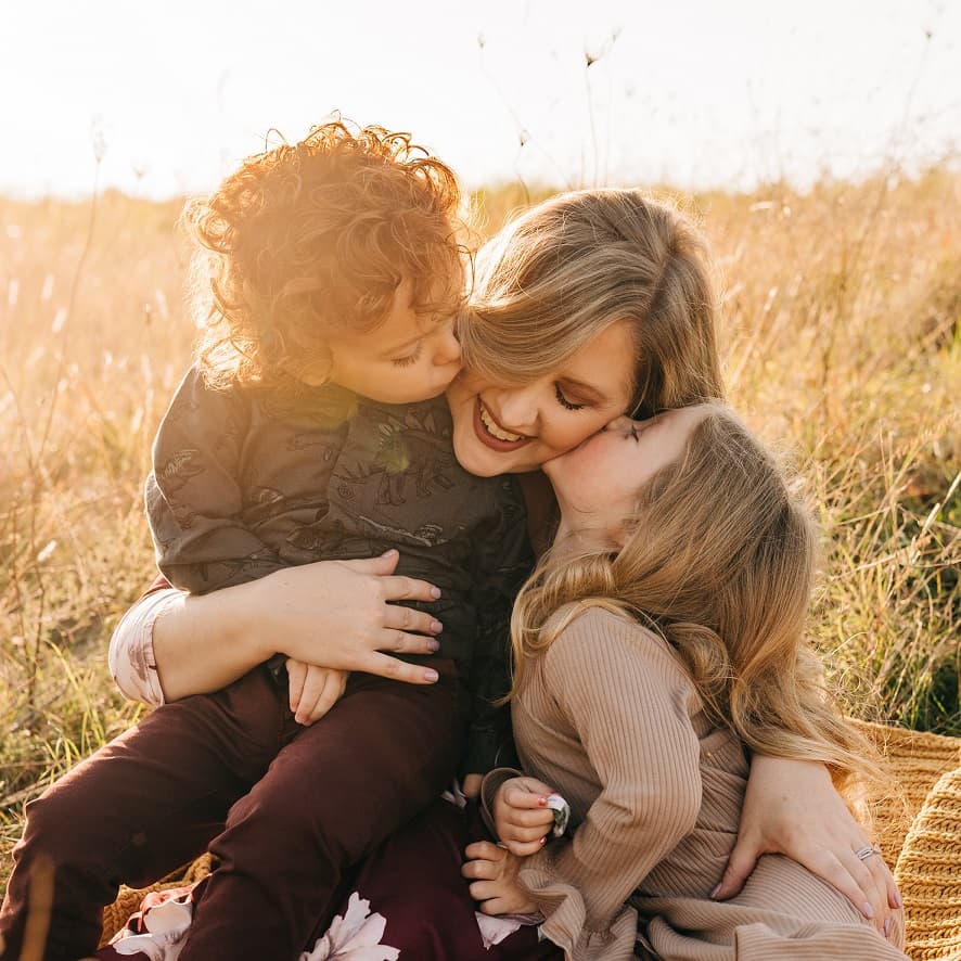 two children kissing their mother sitting in grass