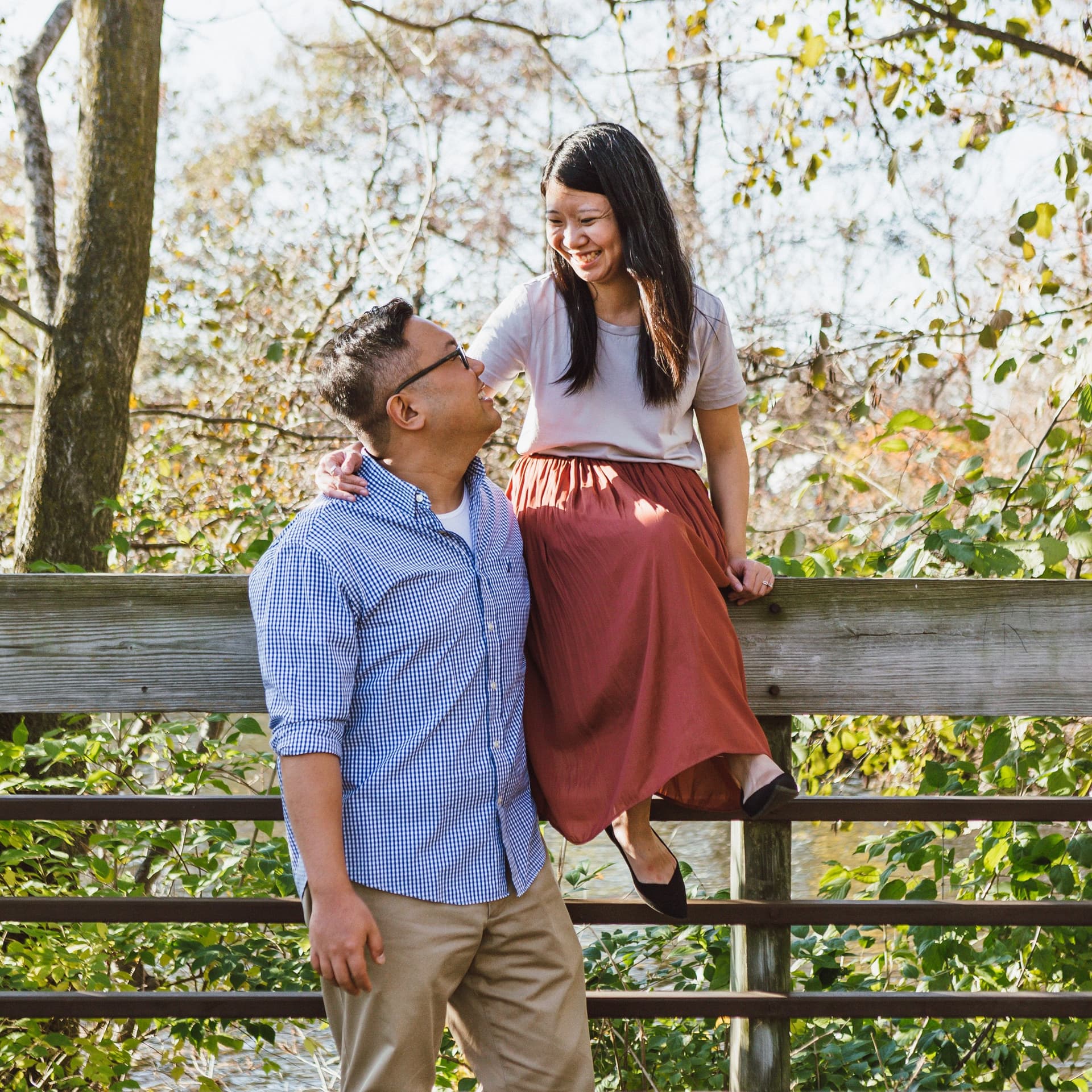 Woman sits on railing while man looks up at her for their free couple photoshoot in a nature preserve