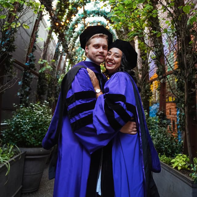 Two graduates in purple regalia embracing and smiling in a garden.