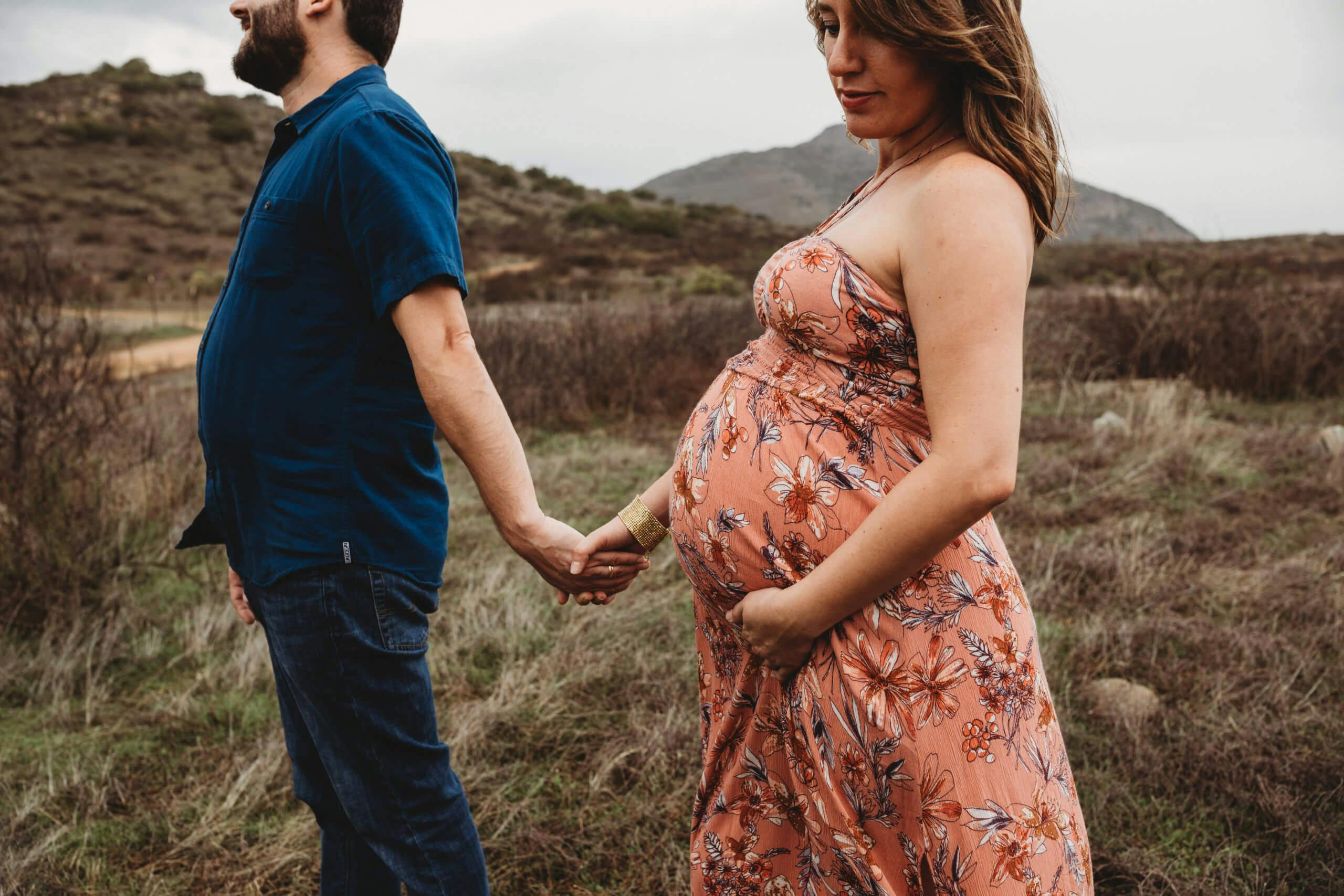 A pregnant woman standing profile with her partner holding her belly in a field