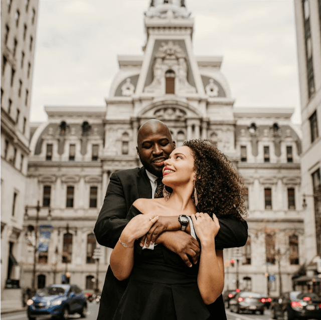 A couple embracing in the foreground with a city hall building in the background and a slightly overcast sky.