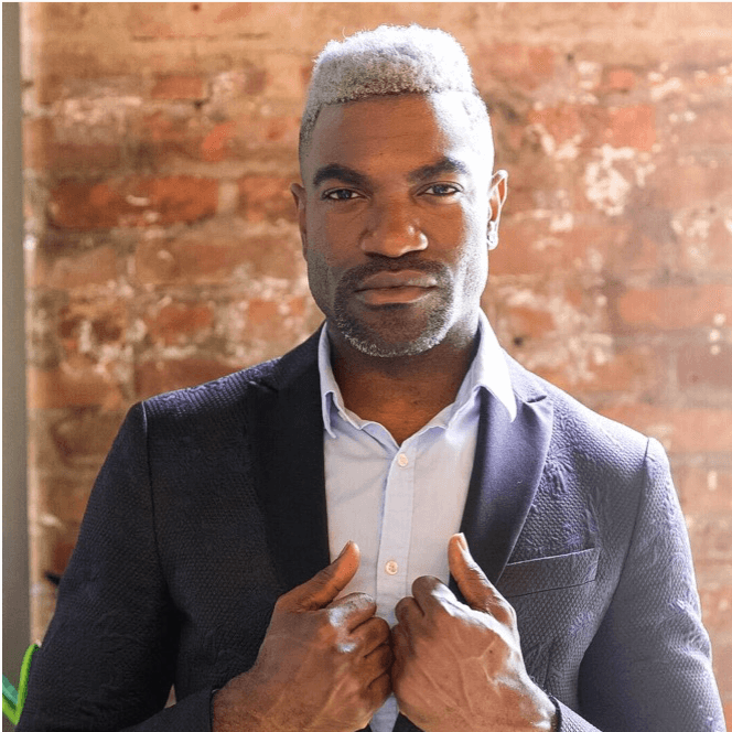 A man with grey hair and a beard, in a suit jacket and a shirt without a tie, staring contemplatively.