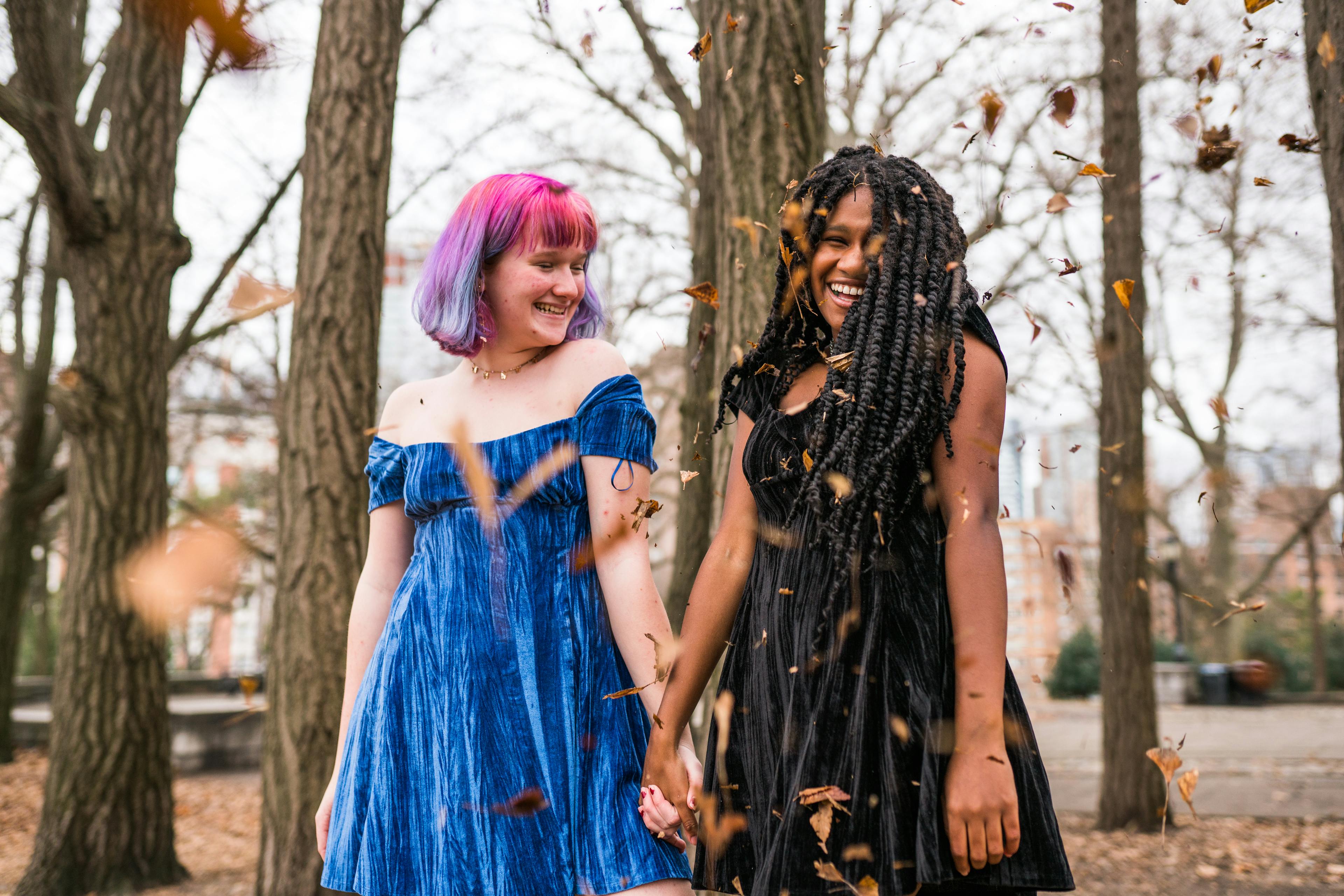 Two friends with colorful hair throwing leaves and laughing in an autumnal park setting