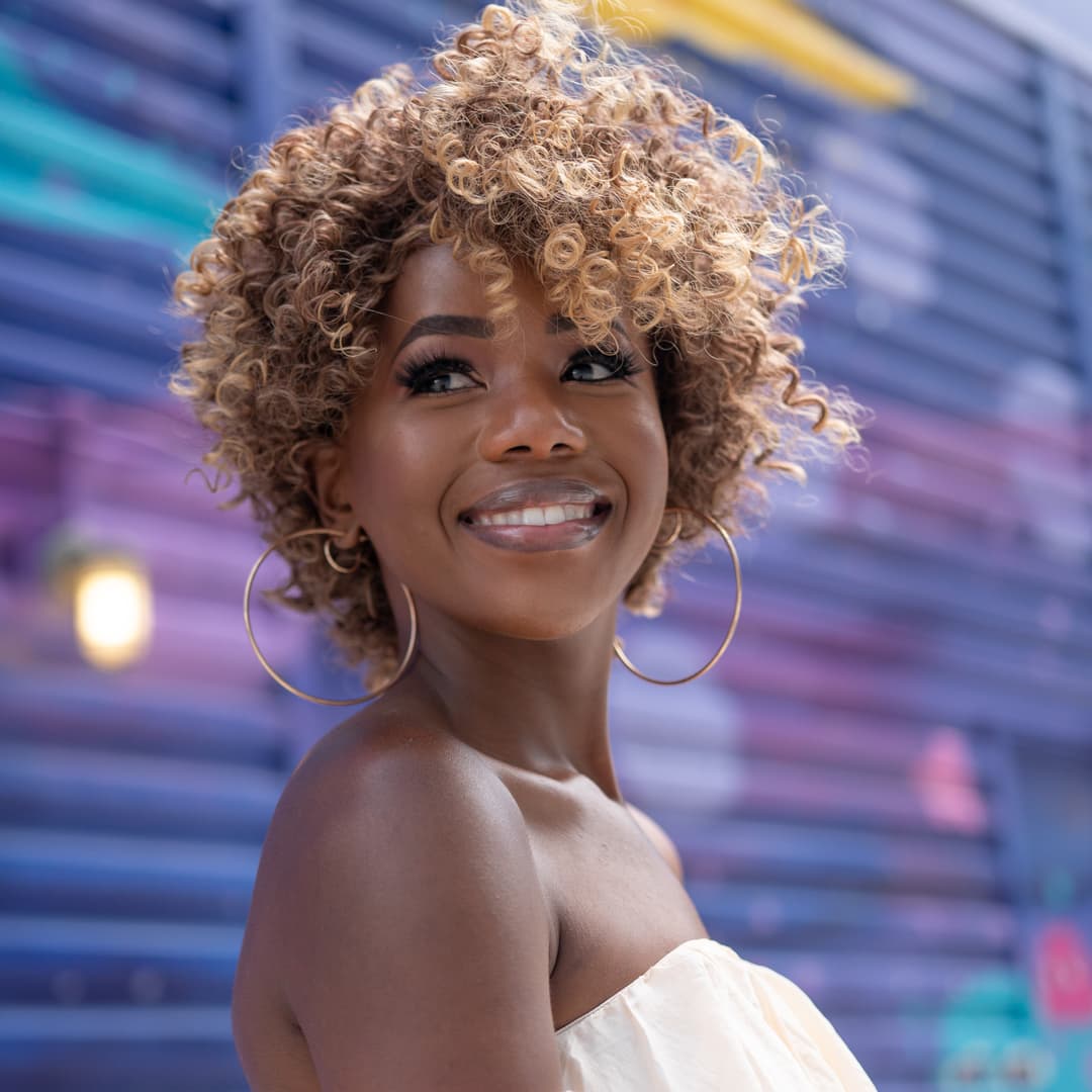 Smiling woman with curly blonde hair posing in natural light during a free Shoott dating profile photography session, colorful outdoor background