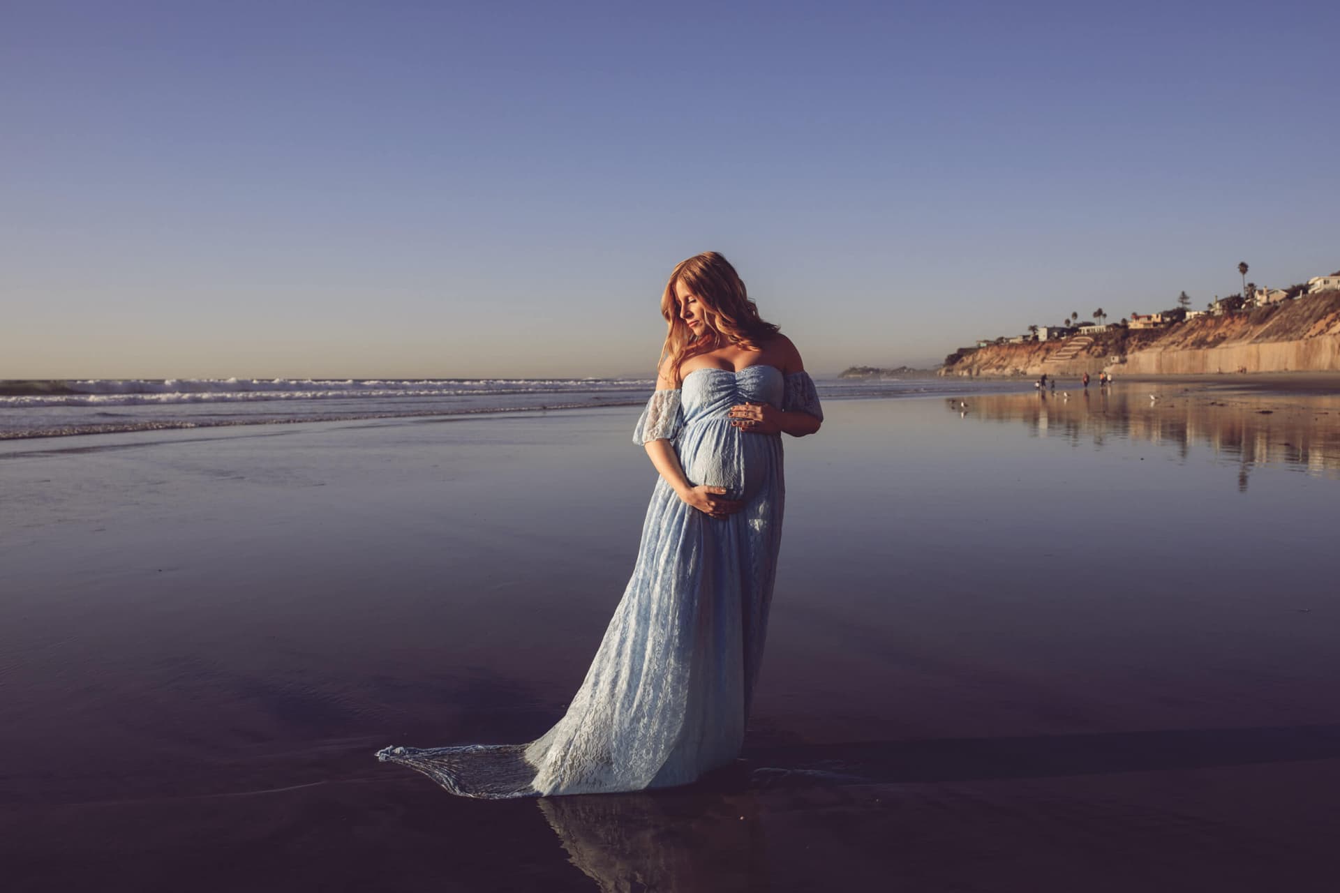Pregnant woman in a blue dress standing on the beach with sunset light