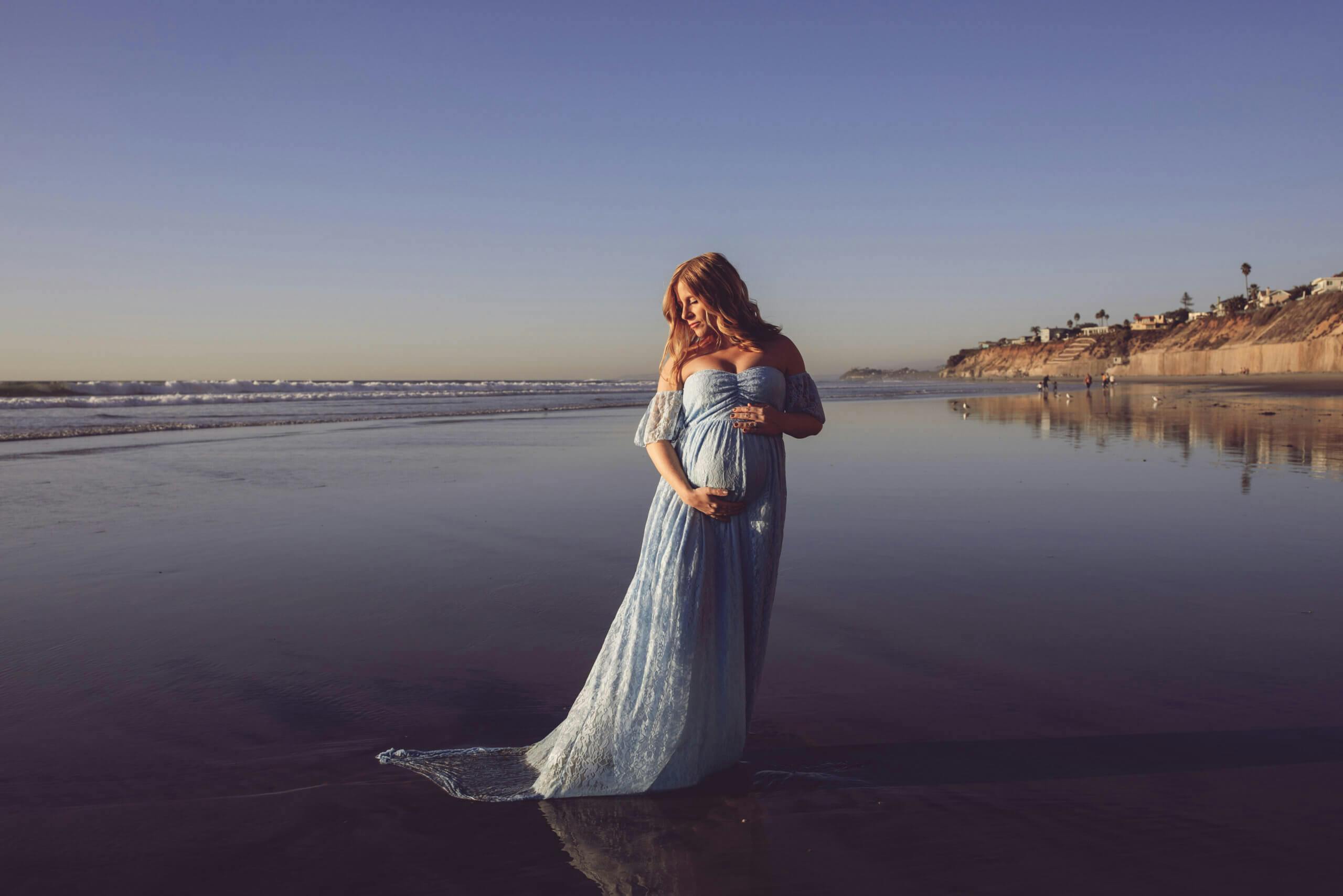 Pregnant woman in a blue dress standing on the beach with sunset light