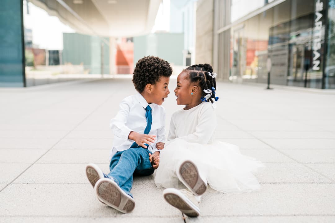 Two joyful children sitting and playfully interacting with each other on a city sidewalk.