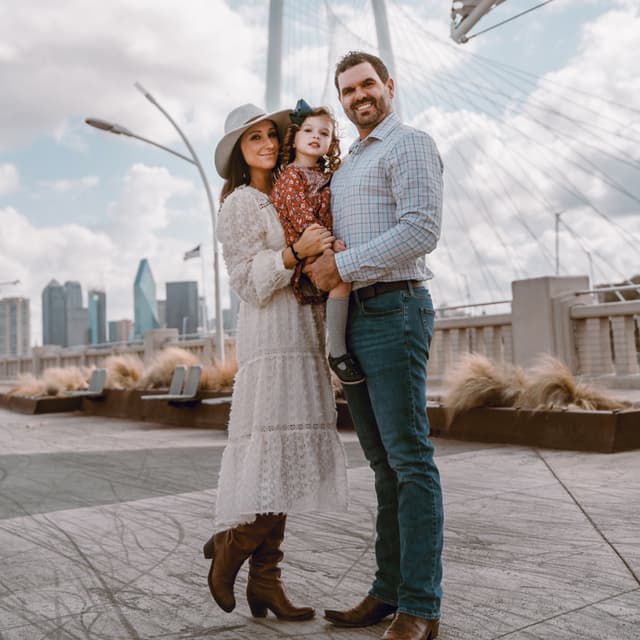 A family of three, with a child in the center, standing on a rooftop with skyscrapers and a bridge in the background.