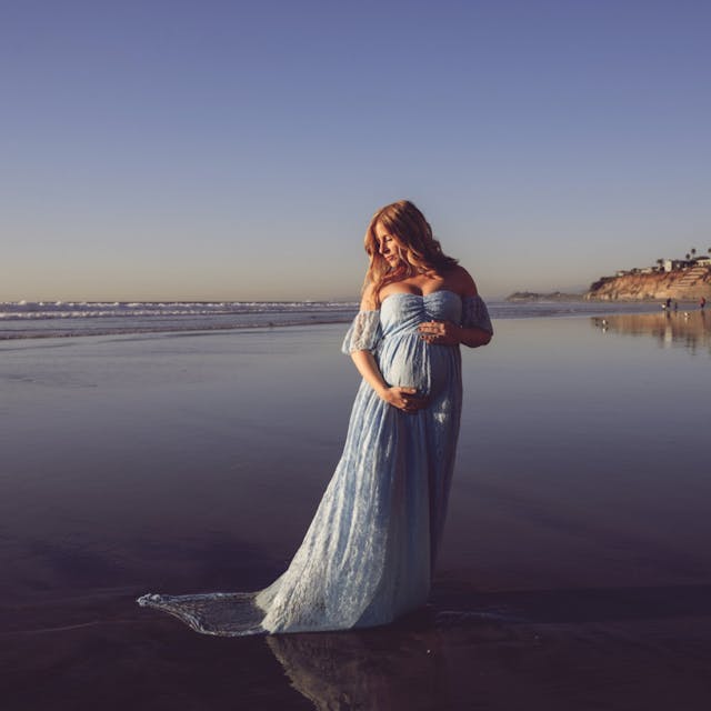 A pregnant woman standing on the beach at sunset, wearing a long blue dress.