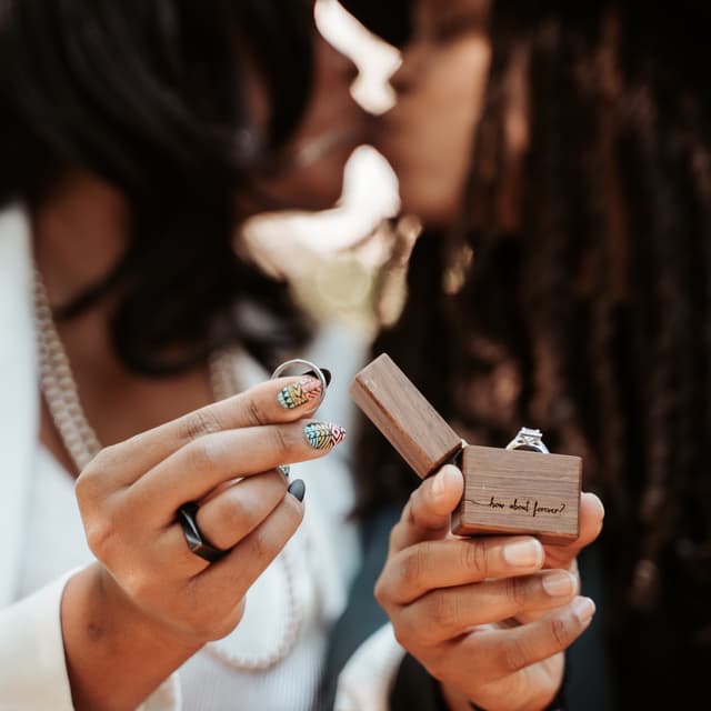 Two people kissing in the background with a focus on a hand holding an open engagement ring box with the phrase "how about forever?"