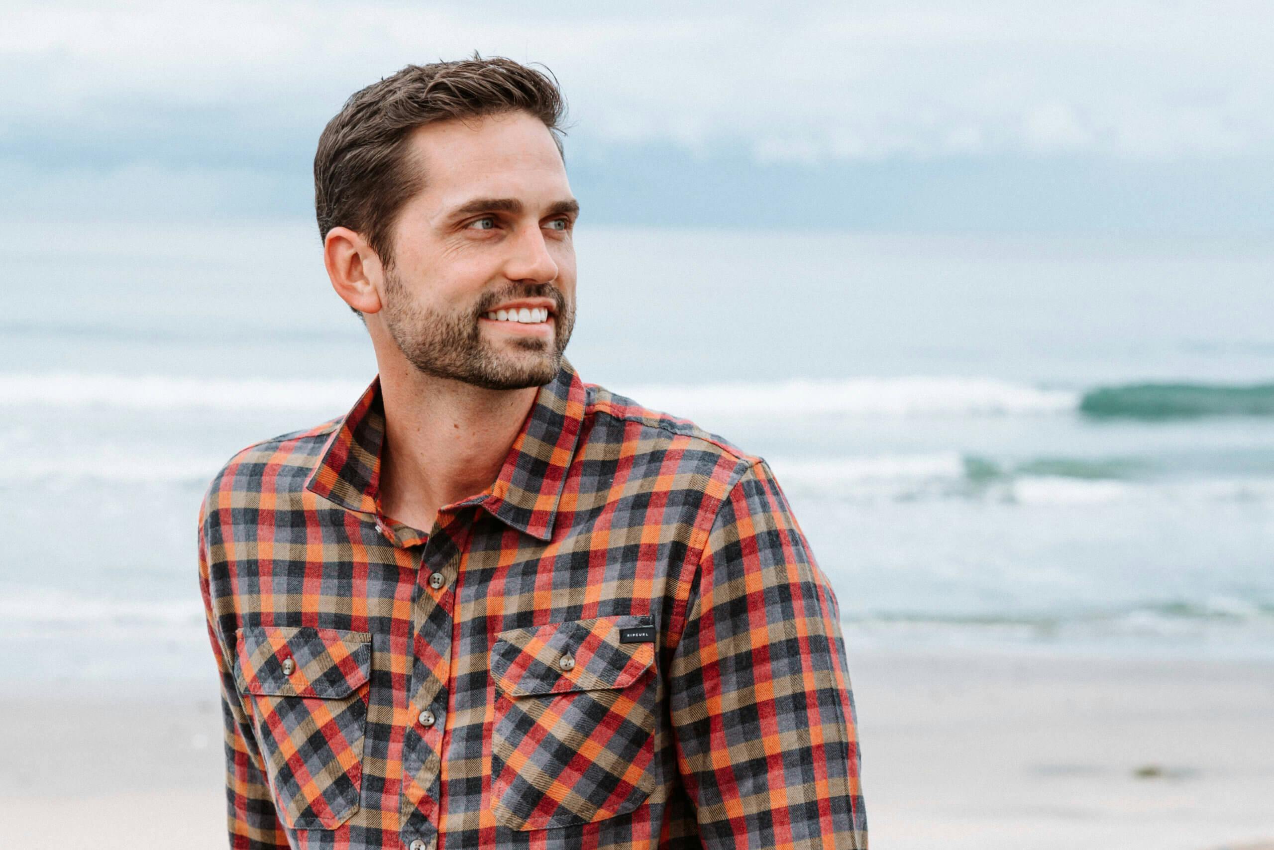 A close-up of a smiling man wearing a plaid shirt with a beach background
