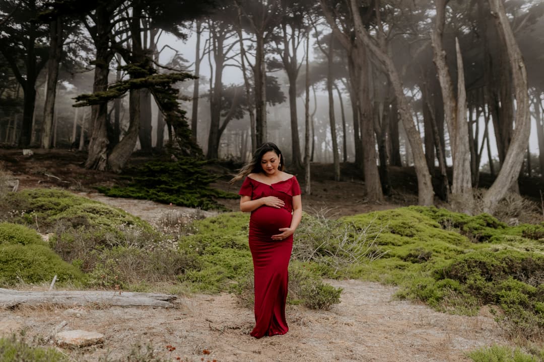 Pregnant woman in a red off-shoulder gown standing on a path through a foggy, wind-swept coastal forest, cradling her belly, captured during a Shoott maternity photoshoot.