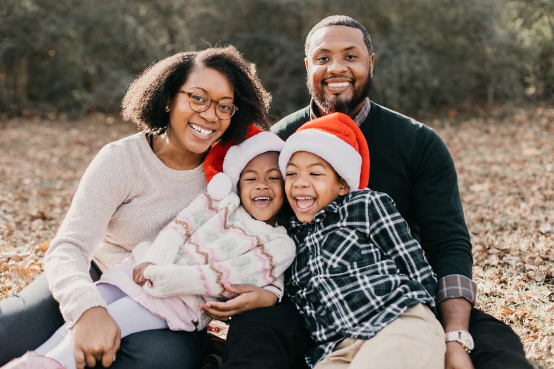 Family of four laughing together outdoors in fall foliage, kids wearing Santa hats, captured during their Shoott holiday session.