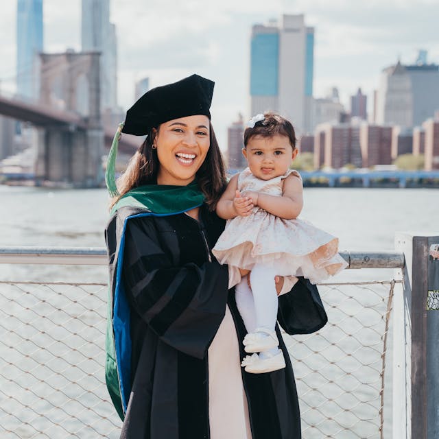 A woman in graduation attire holding a toddler, with a cityscape and bridge in the background.