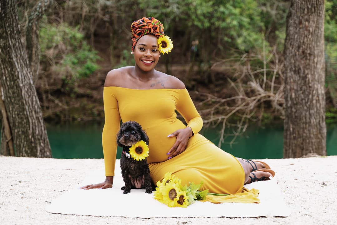 Pregnant woman in a yellow dress sitting with a small black dog surrounded by sunflowers