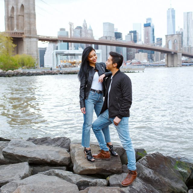 A couple standing on rocks by a river with the Brooklyn Bridge and Manhattan skyline in the background.