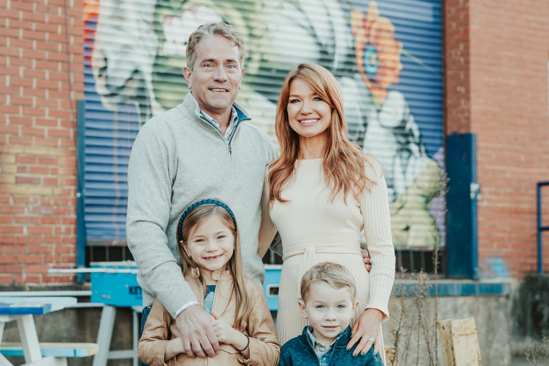 Family of four smiling outdoors in front of a colorful mural during a Shoott mini photoshoot, with mom, dad, daughter, and son posed together against a brick and mural backdrop.
