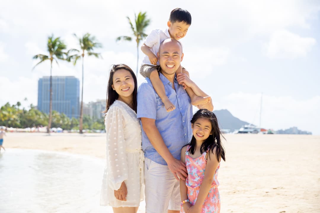 Family of four smiling on a sunny Waikiki beach, with the father carrying his young son on his shoulders and Diamond Head visible in the background, captured during a Shoott family photoshoot.