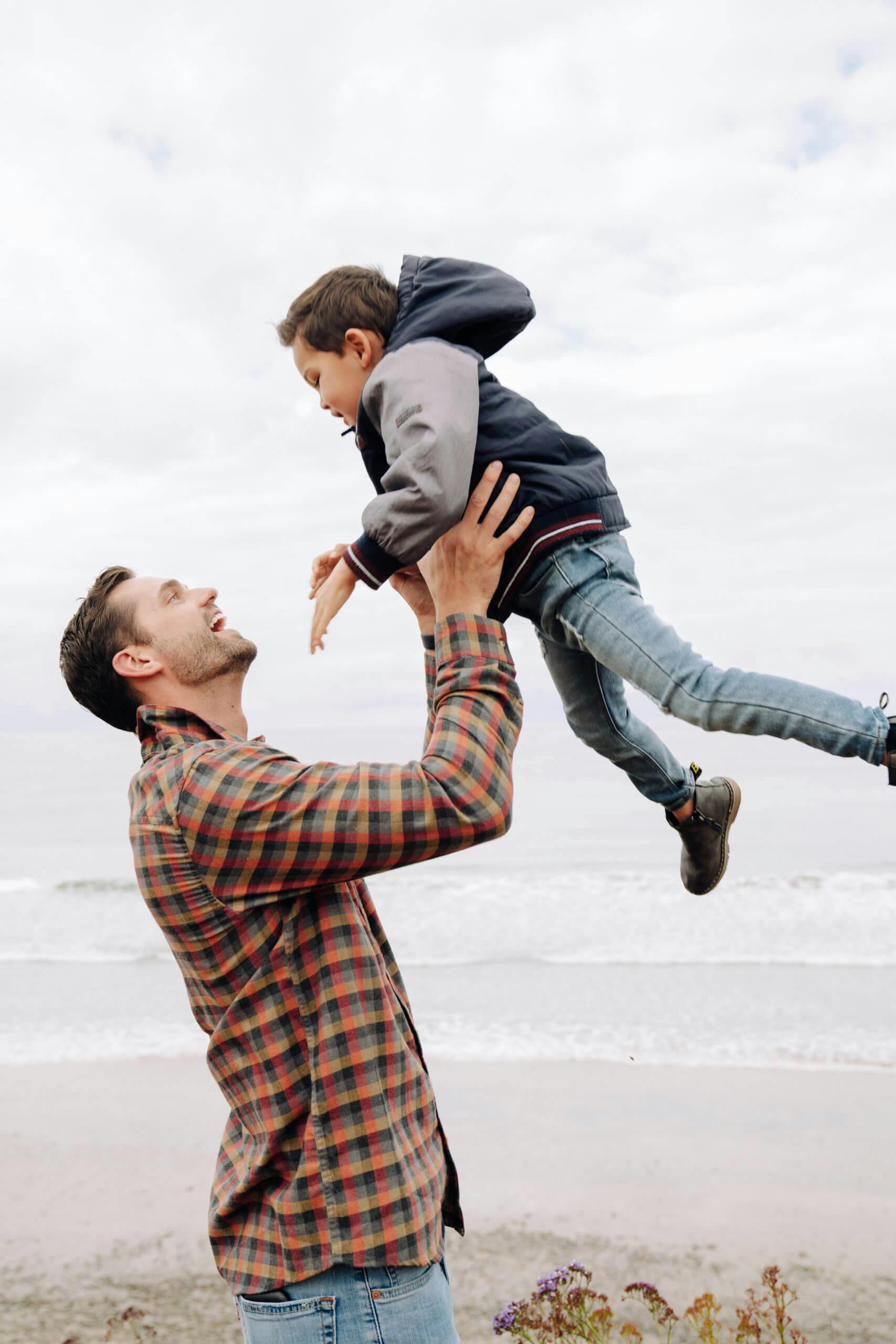 A man lifting a young boy into the air on a beach with overcast sky