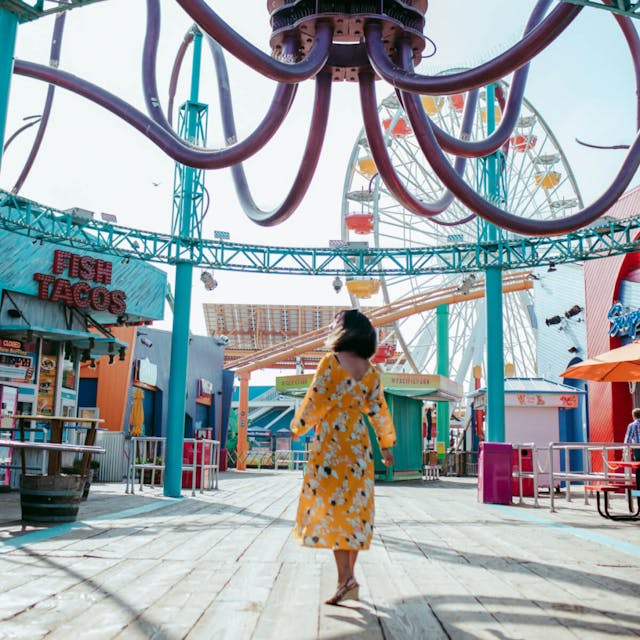 A woman walking through an amusement park with a roller coaster and colorful structures around her.