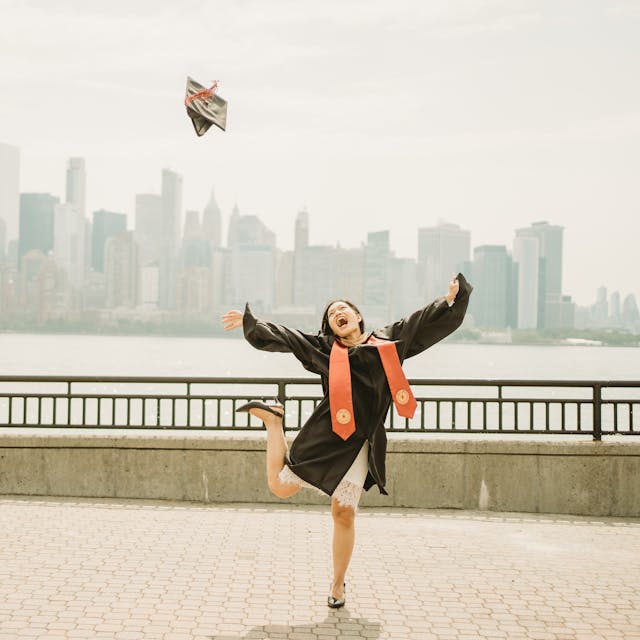 Graduating student in cap and gown joyfully tossing her mortarboard in the air with a city skyline in the background.