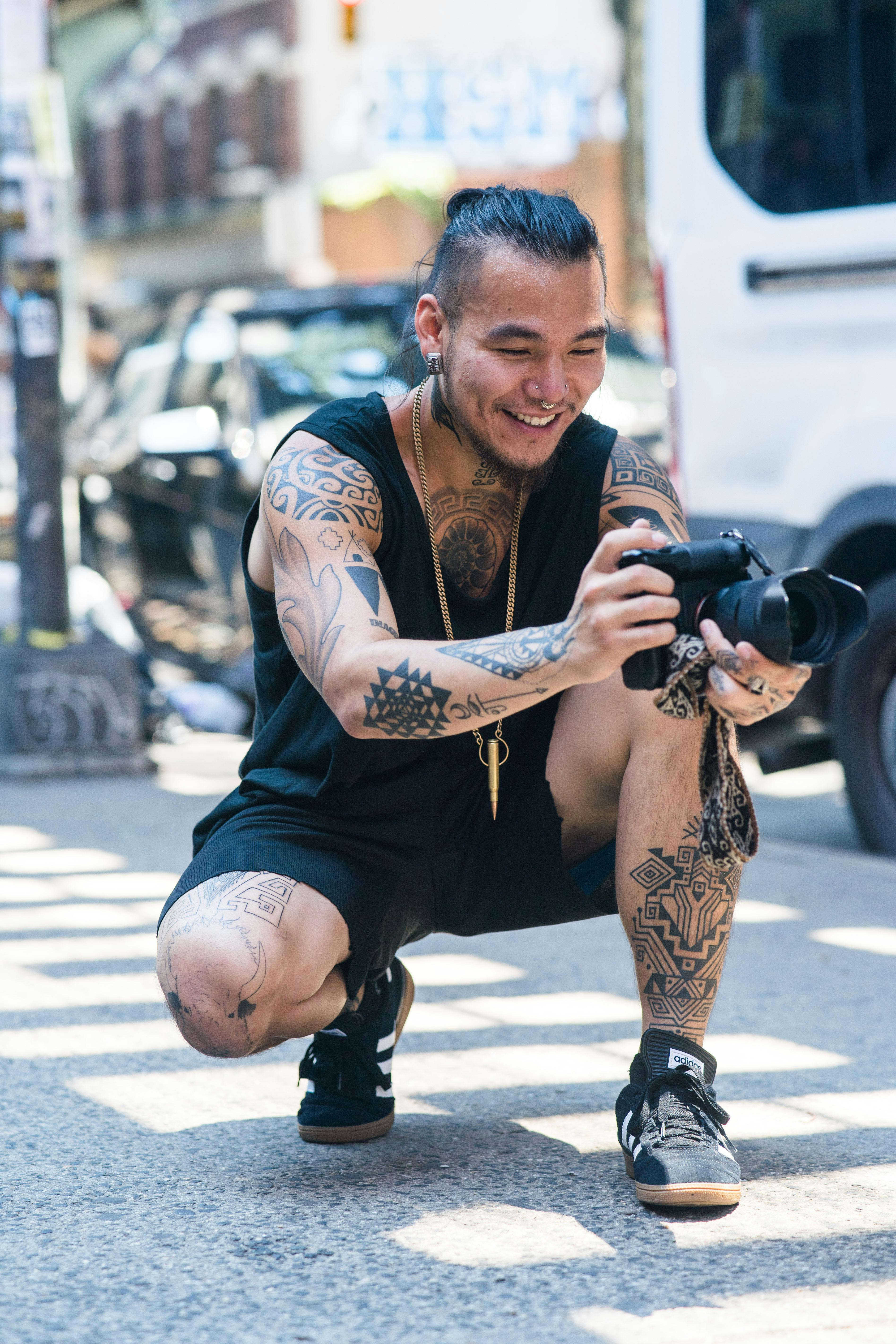 A tattooed photographer crouches with his camera on a city street.