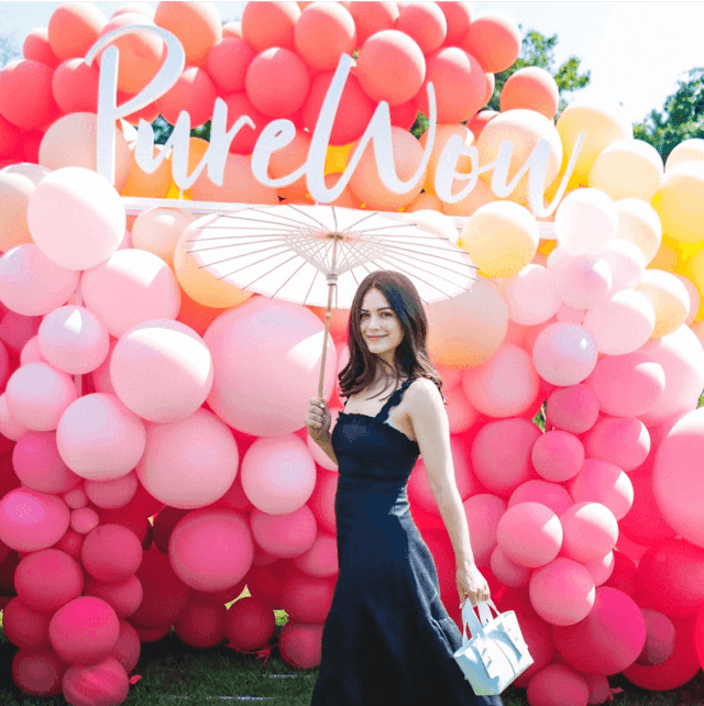 A woman holding an umbrella in front of a wall of pink balloons with the text 'PureWow'.