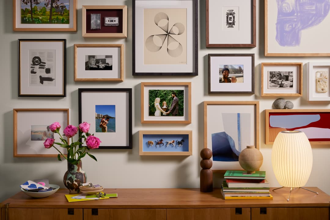 A stylishly decorated room with a gallery wall of framed artwork and photographs, a wooden sideboard, a lit table lamp, books, and a vase with pink flowers.