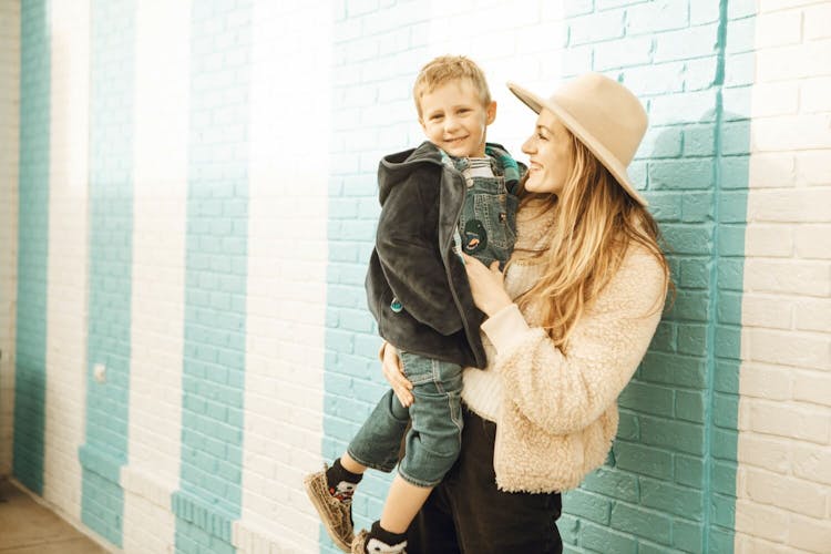 A smiling woman in a hat holding a child against a blue brick wall