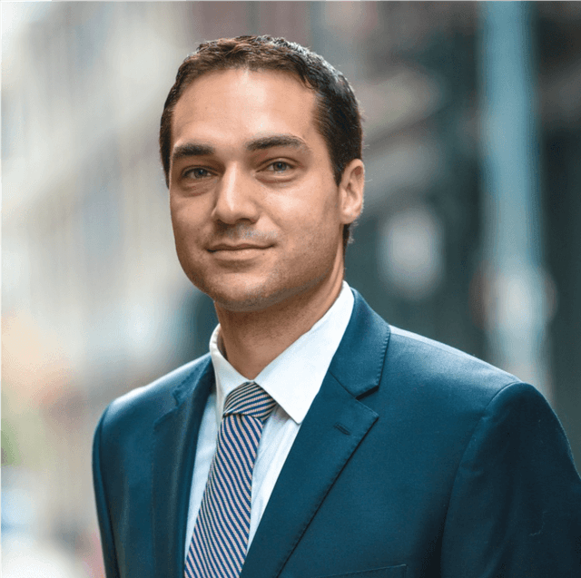 Portrait of a man with dark hair in a suit and tie, standing outdoors with a building blurred in the background.