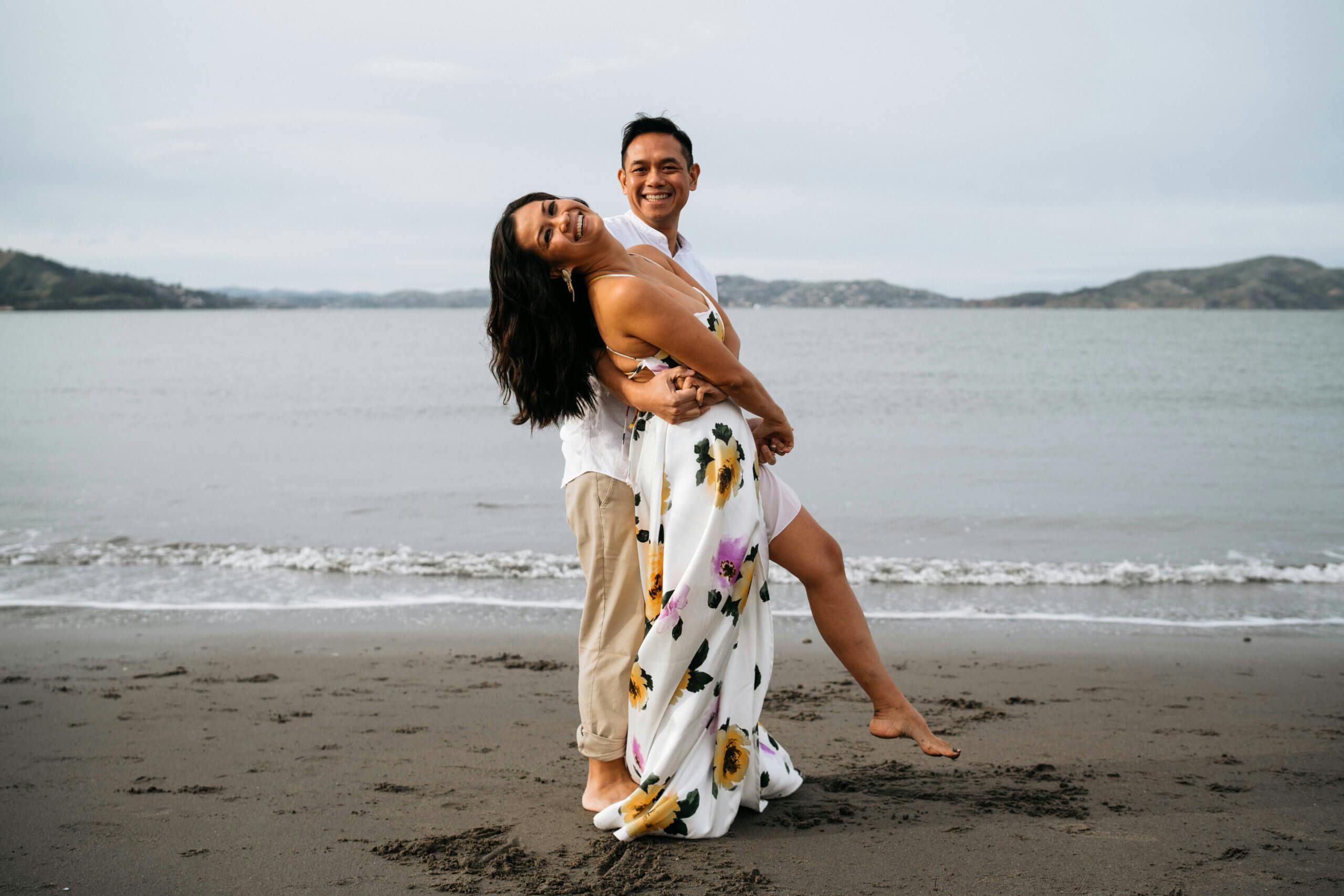 A smiling couple dancing on the beach with hills across the water.