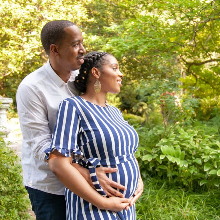 Expectant couple embracing outdoors with the man standing behind the woman, who is cradling her baby bump.