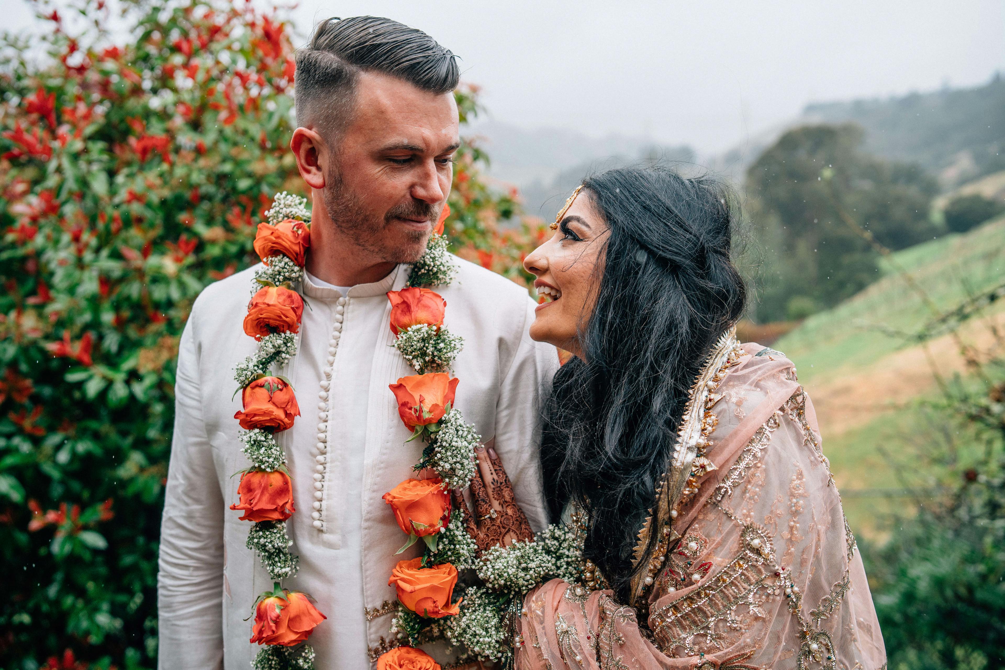 A couple dressed in traditional attire, looking at each other with a backdrop of flowers and fog.