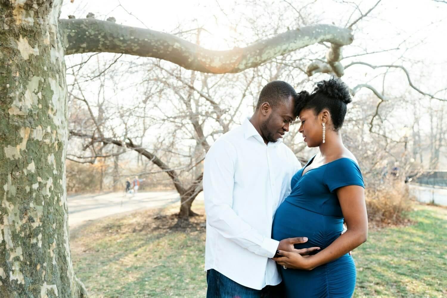 A pregnant woman and her partner touching her belly under a tree