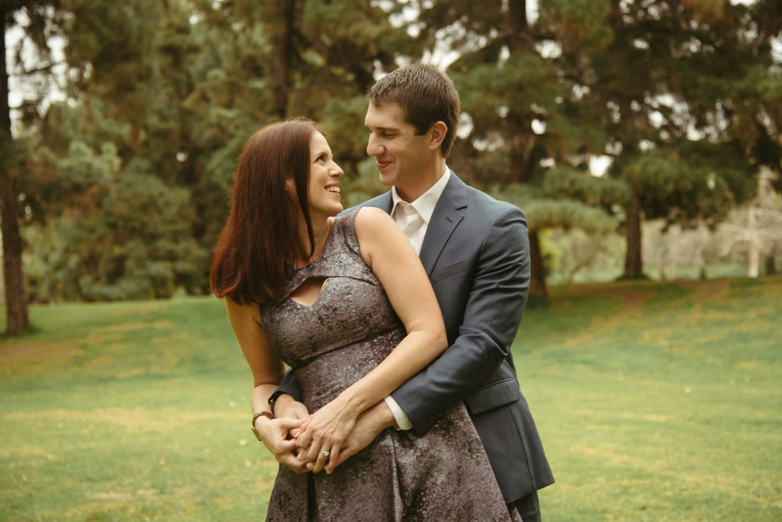 A smiling couple embracing in a park with trees in the background