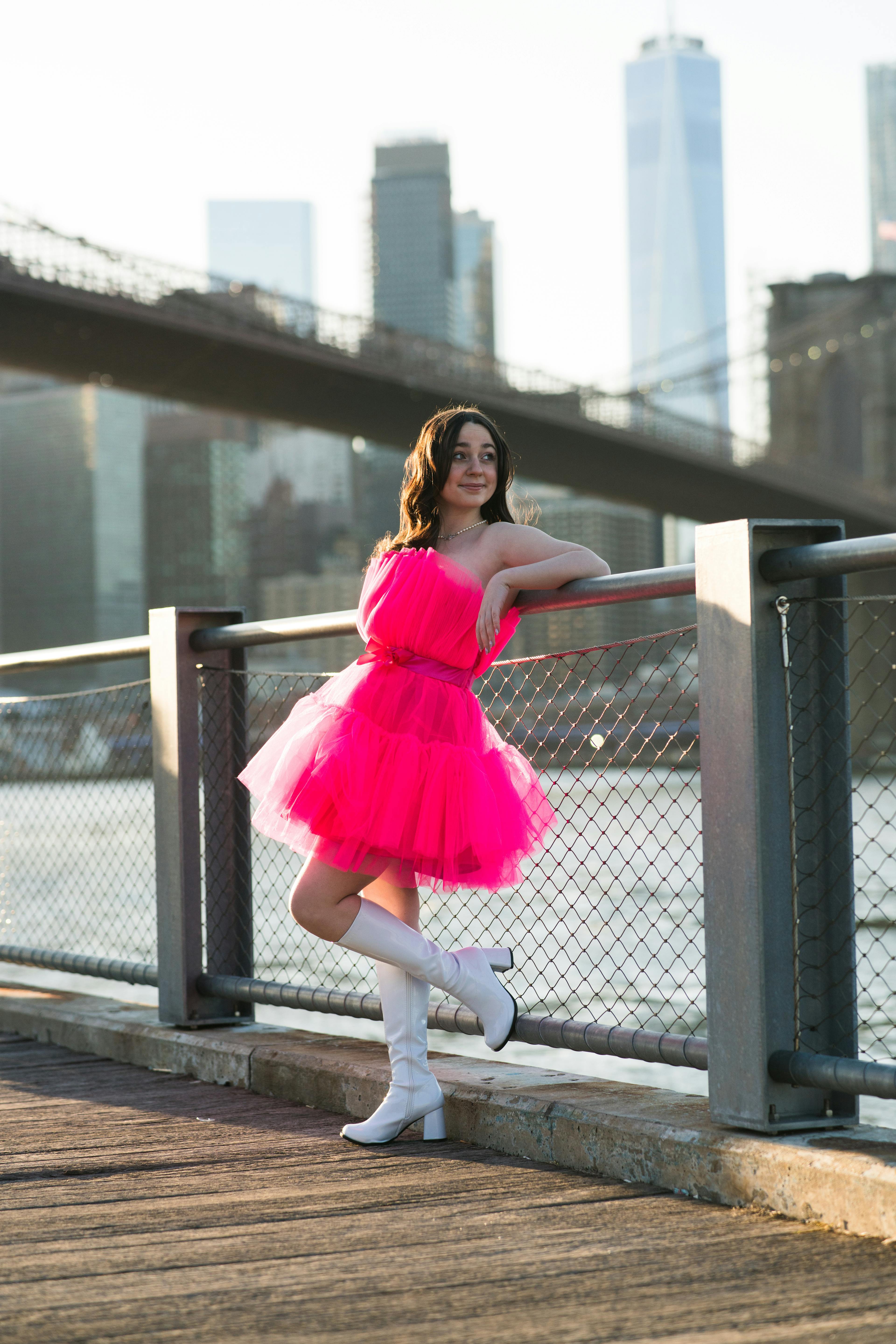 A ballerina in a vibrant pink tutu posing by a railing with the city skyline in the background